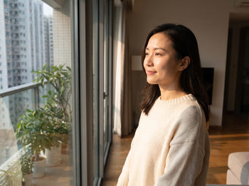 A woman in Hong Kong looking out of an apartment window in autumn light, with calm hydrated skin during the seasonal change